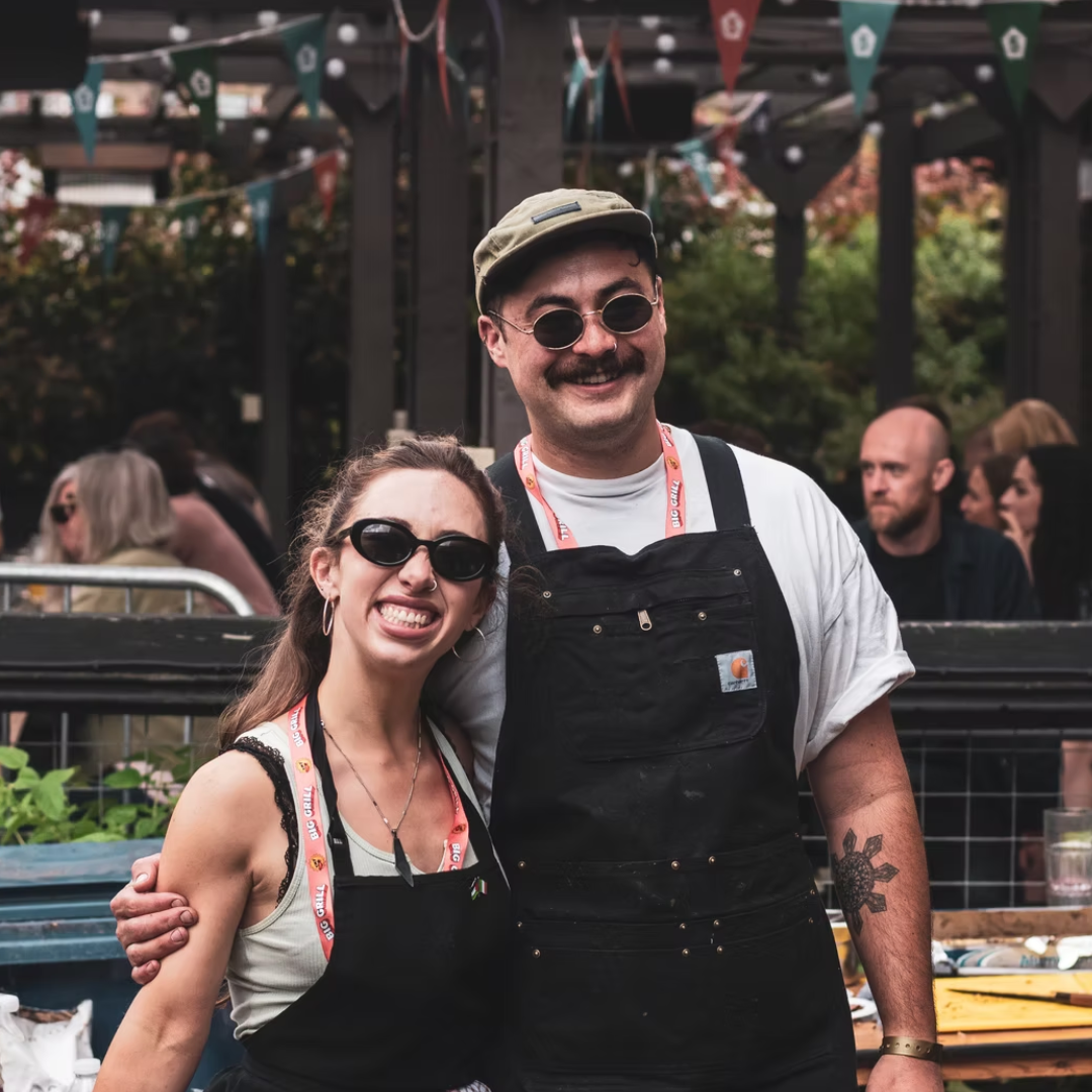 Richie Castillo and Alex O’Neill of Bahay smiling together at a food event, wearing aprons and lanyards, surrounded by people and garden planters.